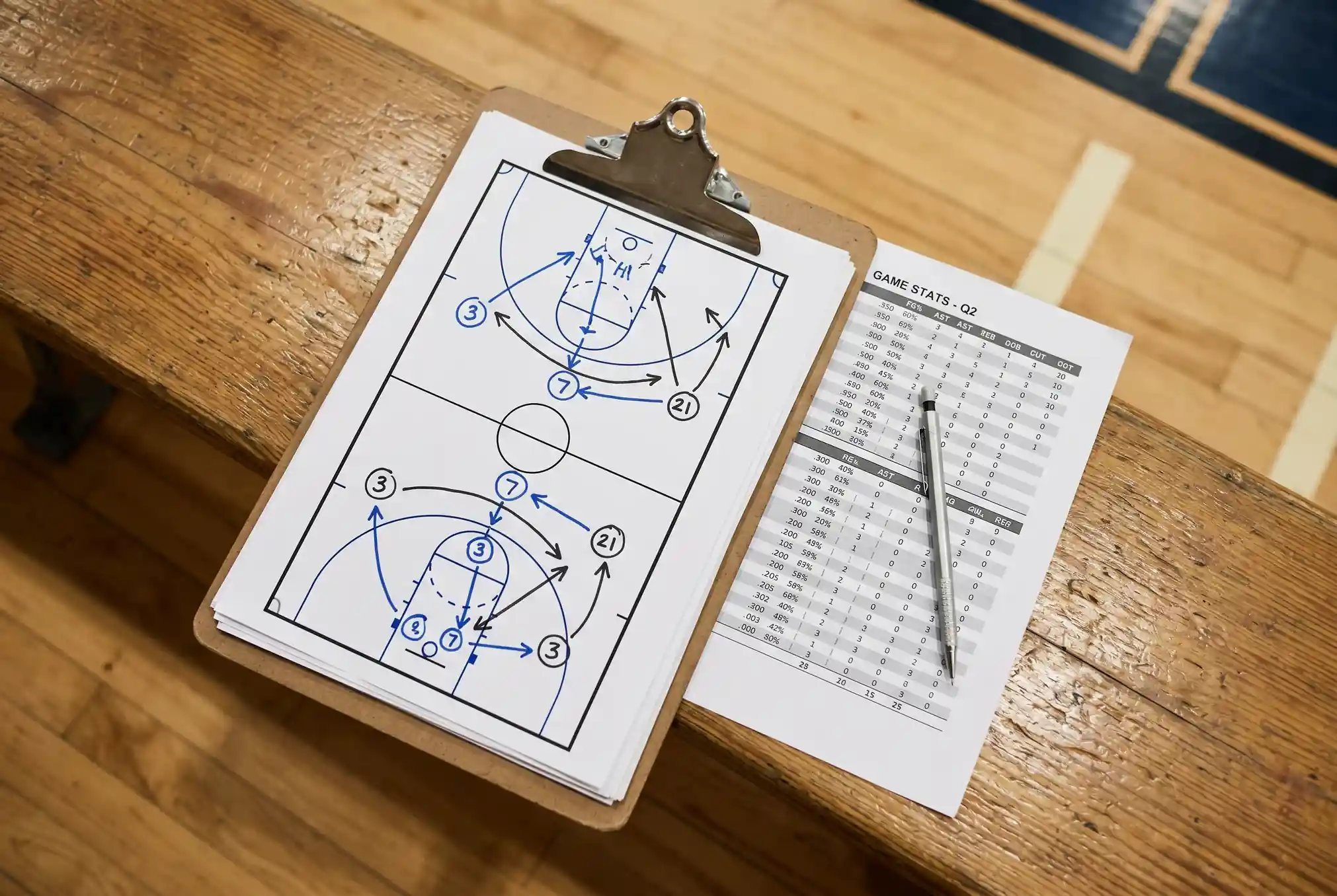 Close-up of a coach's clipboard showing basketball play diagrams beside a printed stat sheet on a bench